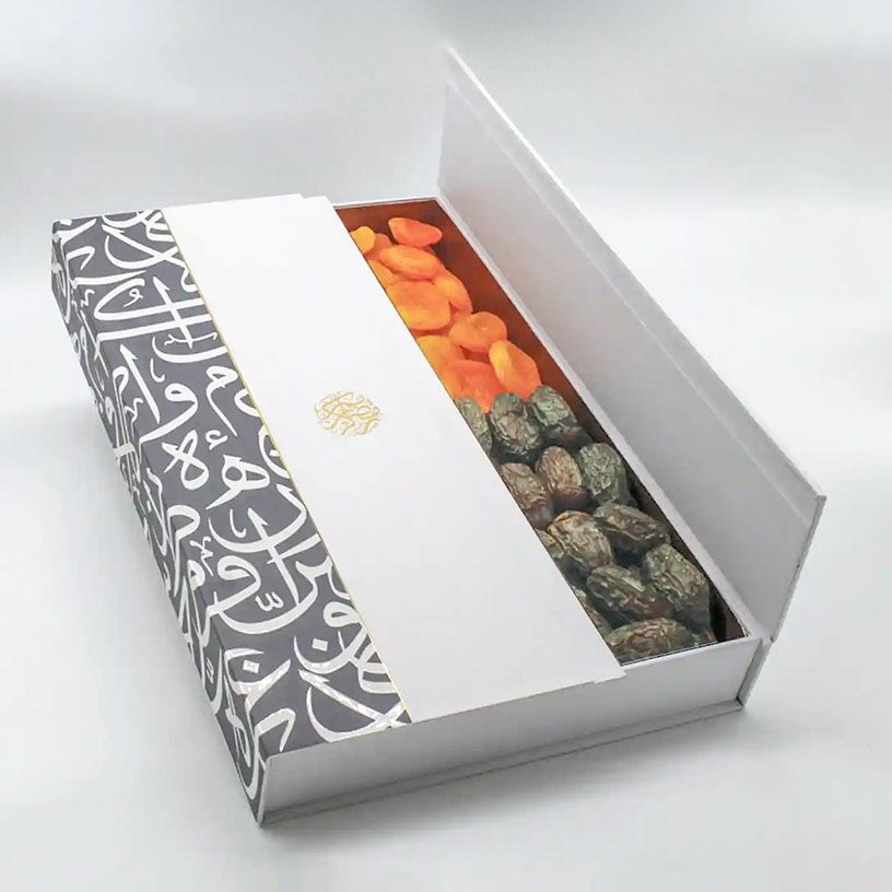 White cardboard gift box adorned with black Arabic calligraphy and a gold geometric accent strip, open to display dried fruits